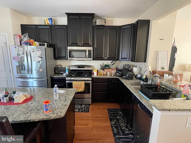 kitchen with light wood finished floors, light stone counters, stainless steel appliances, dark cabinetry, and a sink