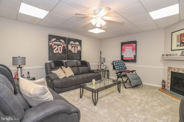 carpeted living room with a stone fireplace, baseboards, ceiling fan, and a drop ceiling