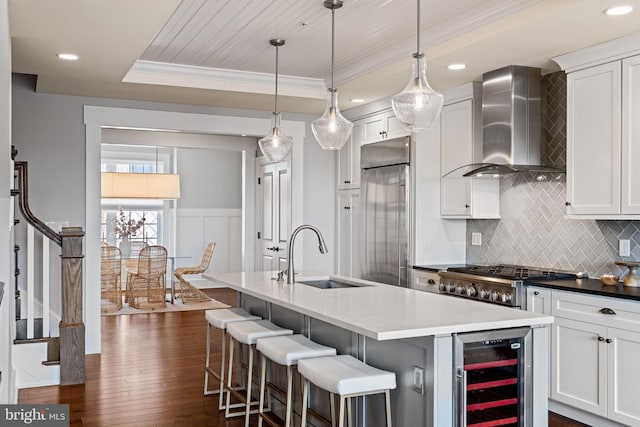 kitchen with wine cooler, a tray ceiling, a sink, stainless steel built in refrigerator, and wall chimney exhaust hood