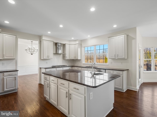 kitchen featuring dark wood finished floors, stainless steel gas cooktop, white cabinets, a kitchen island with sink, and wall chimney exhaust hood