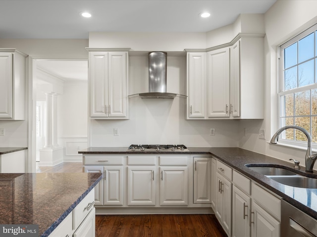 kitchen with decorative columns, wall chimney exhaust hood, appliances with stainless steel finishes, white cabinetry, and a sink