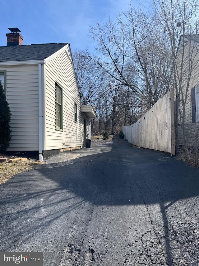 view of property exterior with driveway, a shingled roof, a chimney, and fence