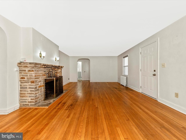 unfurnished living room featuring arched walkways, baseboards, radiator, light wood-style flooring, and a fireplace