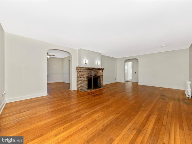 unfurnished living room with arched walkways, baseboards, radiator, light wood-type flooring, and a brick fireplace