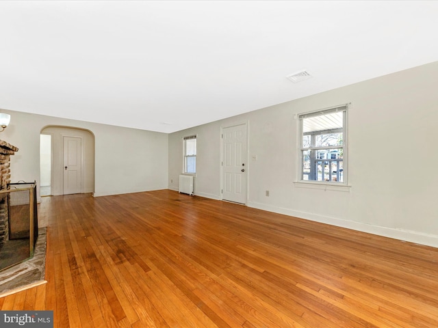 living area featuring arched walkways, baseboards, radiator heating unit, light wood-style flooring, and a stone fireplace