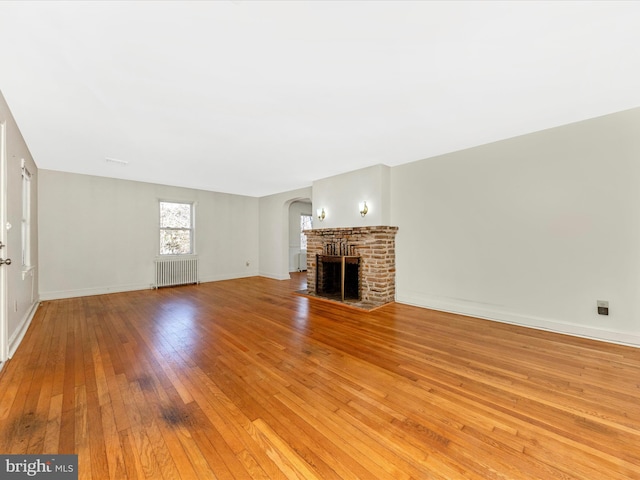 unfurnished living room featuring arched walkways, a fireplace, radiator, light wood-style floors, and baseboards