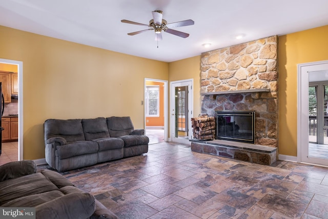 living room with a ceiling fan, baseboards, stone tile flooring, and a stone fireplace