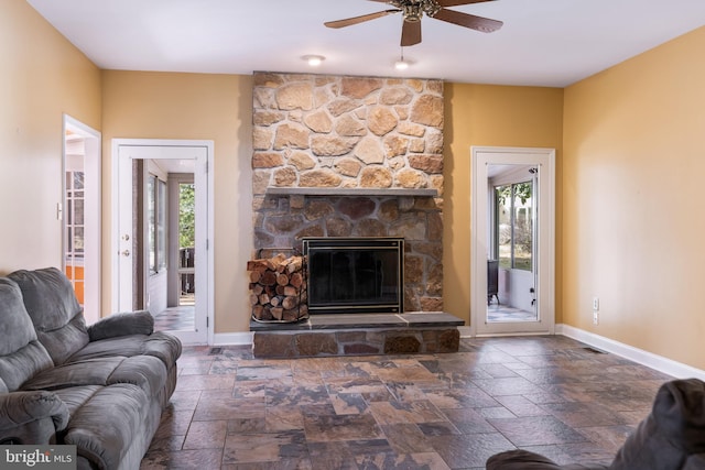 living area featuring stone tile flooring, ceiling fan, a stone fireplace, and baseboards