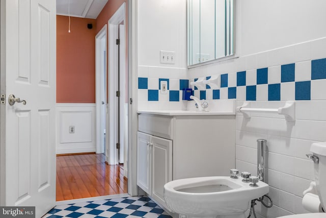 half bathroom with wainscoting, toilet, vanity, a bidet, and tile walls