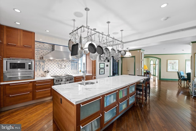 kitchen with stainless steel appliances, a sink, brown cabinets, a warming drawer, and wall chimney exhaust hood