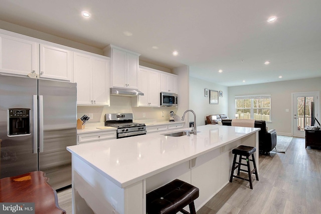 kitchen featuring a center island with sink, stainless steel appliances, light wood-style floors, a sink, and under cabinet range hood