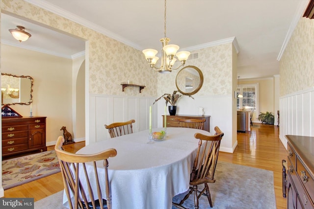 dining space with wallpapered walls, crown molding, light wood-style floors, and a wainscoted wall