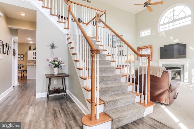 stairway featuring baseboards, a ceiling fan, wood finished floors, a high ceiling, and a fireplace