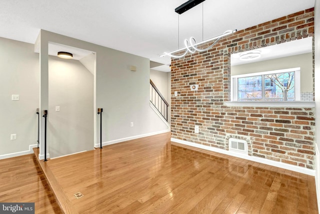 unfurnished living room with brick wall, wood finished floors, visible vents, baseboards, and stairway