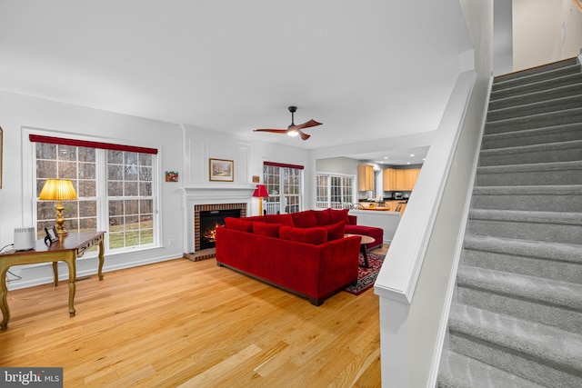 living room featuring a brick fireplace, light wood-style flooring, stairs, and a ceiling fan