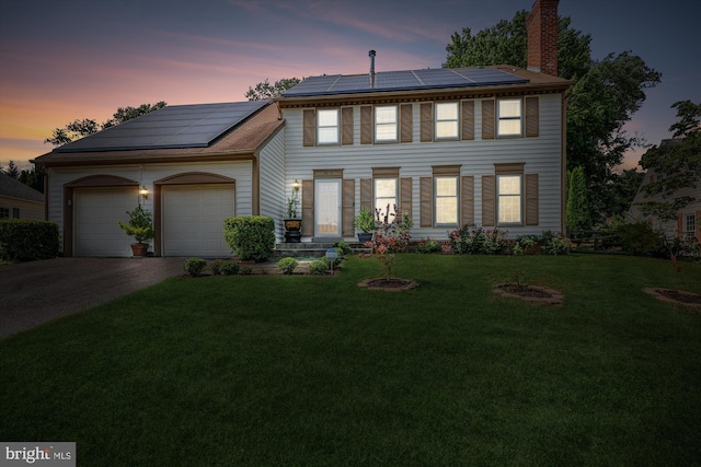 colonial home featuring a garage, a yard, a chimney, and roof mounted solar panels