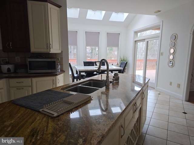 kitchen featuring a sink, stainless steel microwave, dark stone counters, light tile patterned floors, and decorative backsplash