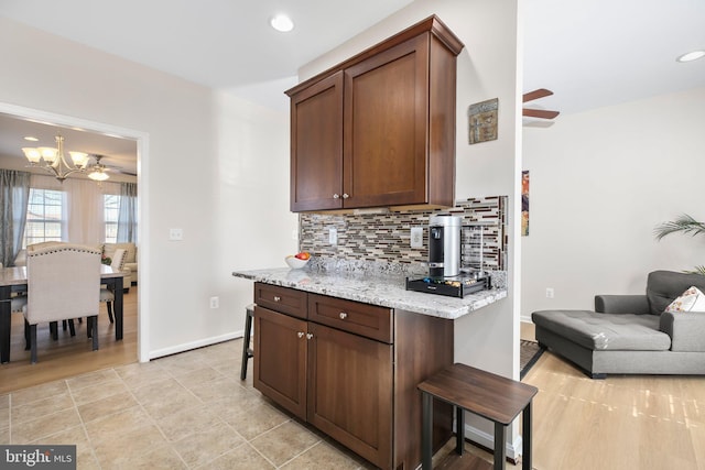 kitchen featuring light stone counters, backsplash, ceiling fan with notable chandelier, and baseboards