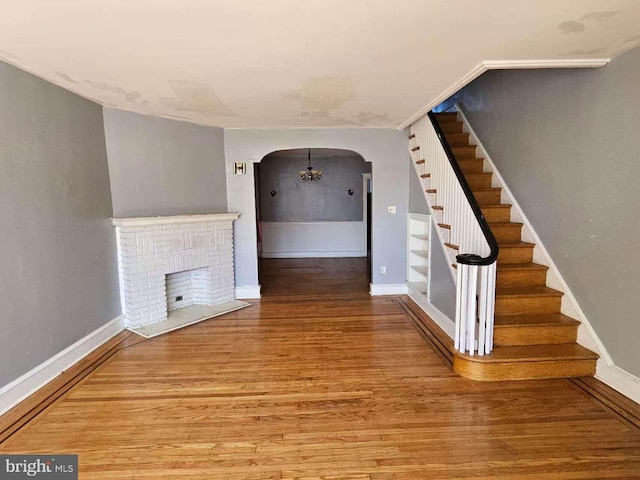 unfurnished living room featuring baseboards, arched walkways, stairway, wood finished floors, and a brick fireplace