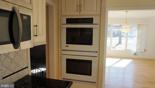 kitchen with white double oven, stainless steel microwave, wood finished floors, dark stone countertops, and a notable chandelier