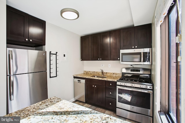 kitchen featuring light stone counters, dark brown cabinetry, wood finished floors, a sink, and appliances with stainless steel finishes