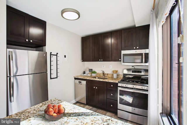 kitchen with light stone countertops, appliances with stainless steel finishes, a sink, dark brown cabinets, and wood finished floors