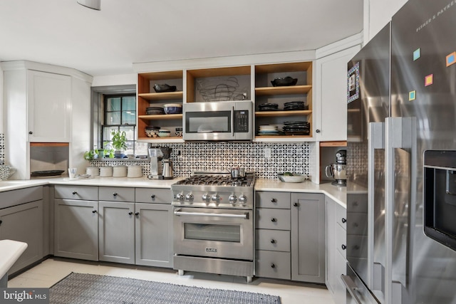 kitchen with gray cabinetry, stainless steel appliances, light countertops, decorative backsplash, and open shelves