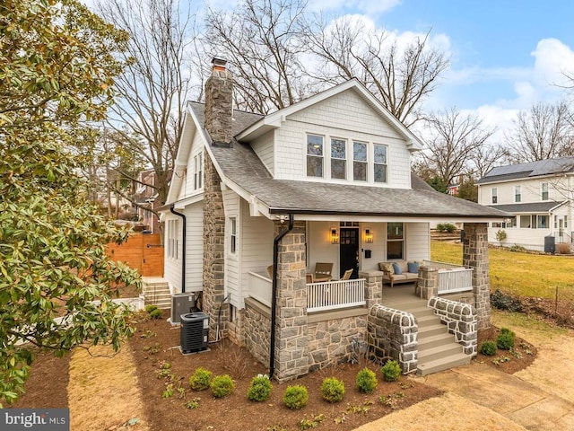 view of front of house featuring a shingled roof, stone siding, a chimney, cooling unit, and a porch