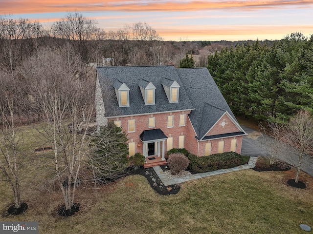 view of front of house featuring a shingled roof, brick siding, a wooded view, and a front lawn