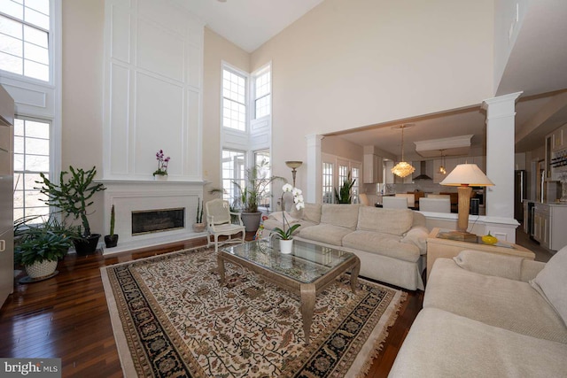 living area with dark wood-type flooring, a glass covered fireplace, a towering ceiling, and ornate columns