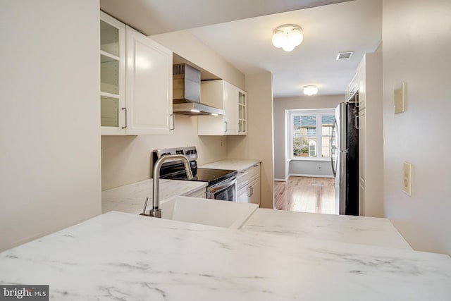 kitchen featuring stainless steel appliances, glass insert cabinets, light wood finished floors, and wall chimney exhaust hood