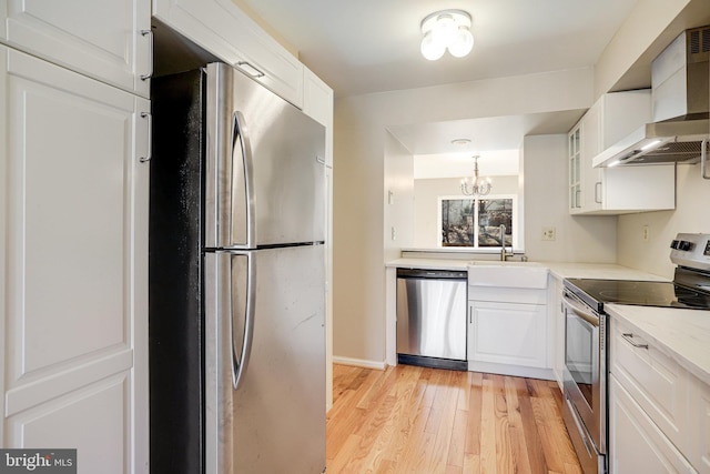 kitchen featuring appliances with stainless steel finishes, glass insert cabinets, white cabinets, wall chimney range hood, and light wood-type flooring