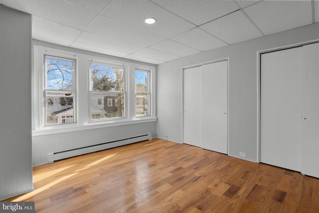 unfurnished bedroom with multiple closets, a drop ceiling, baseboard heating, and wood finished floors