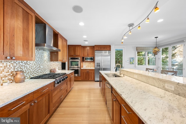 kitchen with light stone counters, brown cabinetry, a sink, wall chimney range hood, and built in appliances