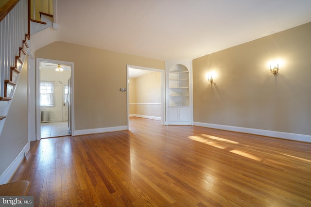 unfurnished living room featuring built in shelves, radiator, hardwood / wood-style floors, baseboards, and stairs