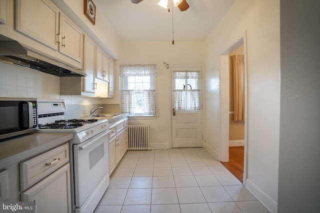 kitchen with white gas stove, stainless steel microwave, radiator heating unit, light tile patterned flooring, and under cabinet range hood