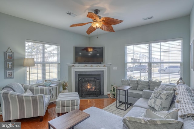 living area featuring a warm lit fireplace, ceiling fan, visible vents, and wood finished floors