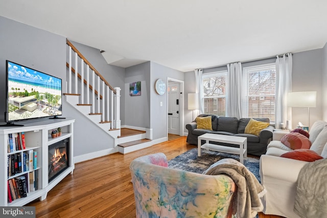 living area featuring stairs, a glass covered fireplace, baseboards, and wood finished floors