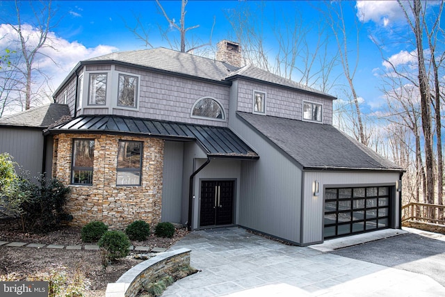 view of front of house featuring driveway, a standing seam roof, an attached garage, a chimney, and metal roof