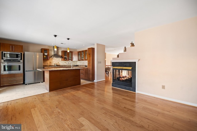 kitchen featuring a center island with sink, wall chimney exhaust hood, appliances with stainless steel finishes, a sink, and a multi sided fireplace