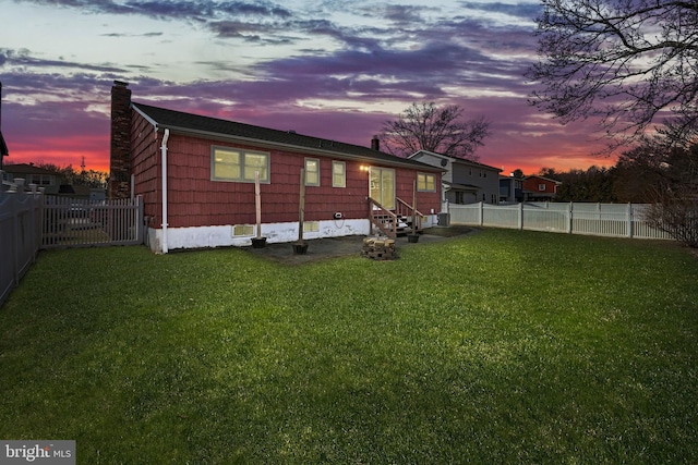 back of house at dusk featuring entry steps, a fenced backyard, a chimney, and a lawn