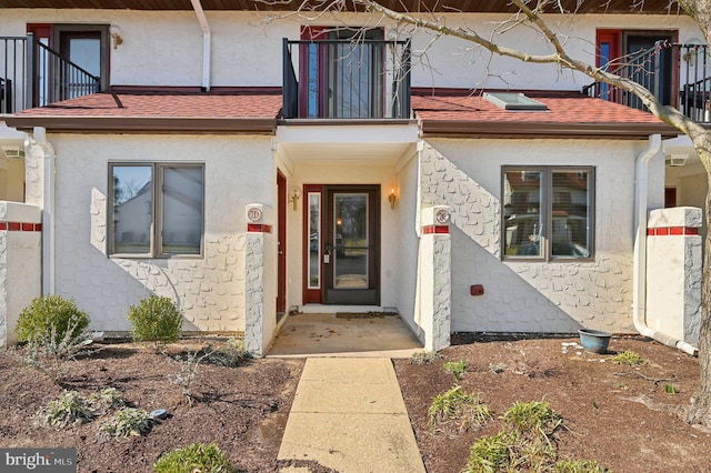 view of exterior entry with stucco siding, a balcony, and a shingled roof