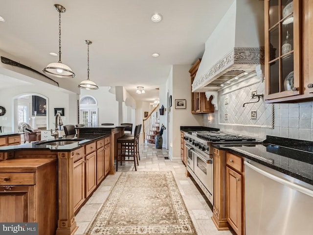 kitchen with custom exhaust hood, backsplash, appliances with stainless steel finishes, brown cabinetry, and a sink