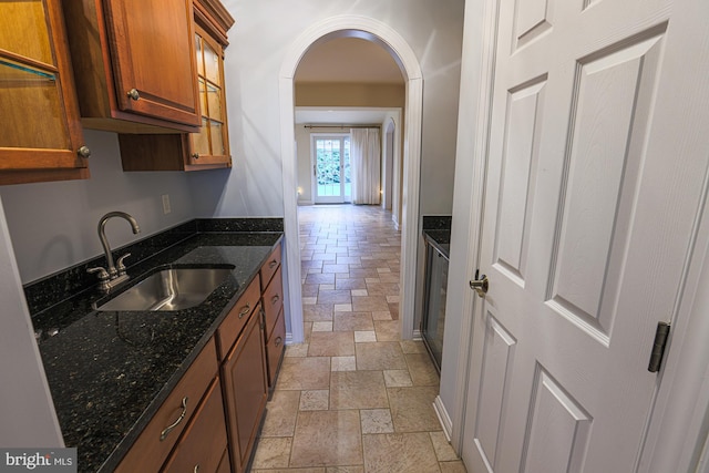 kitchen featuring arched walkways, a sink, brown cabinetry, dark stone countertops, and stone tile flooring