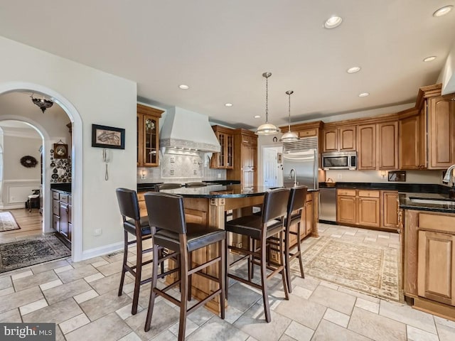 kitchen with a kitchen island with sink, stainless steel appliances, a sink, custom exhaust hood, and a kitchen bar