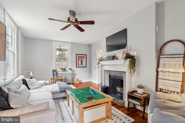 living area with radiator, a fireplace with raised hearth, ceiling fan, wood finished floors, and baseboards