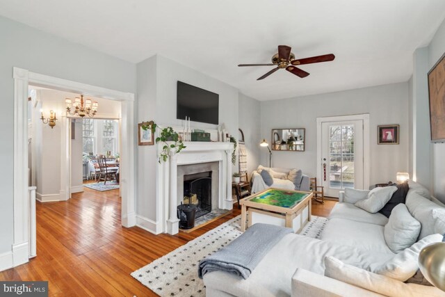 living room featuring light wood-style floors, baseboards, a fireplace with flush hearth, and ceiling fan with notable chandelier