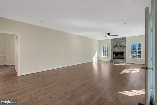 unfurnished living room featuring dark wood finished floors, a stone fireplace, baseboards, and ceiling fan