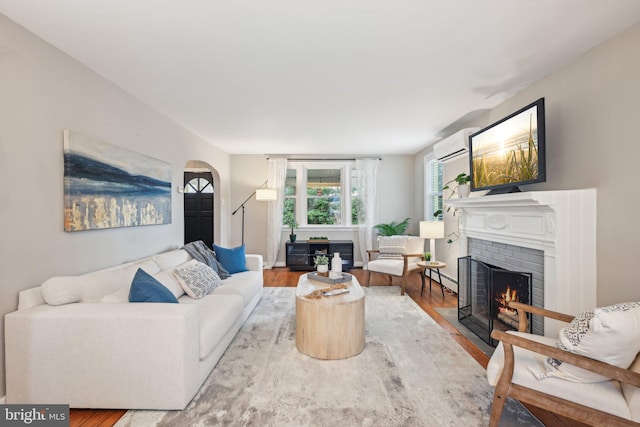 living room with arched walkways, baseboard heating, a brick fireplace, an AC wall unit, and wood finished floors