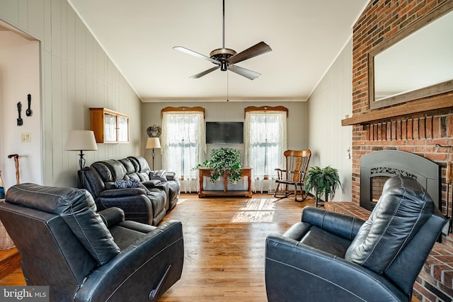 living room with a ceiling fan, ornamental molding, wood finished floors, vaulted ceiling, and a brick fireplace